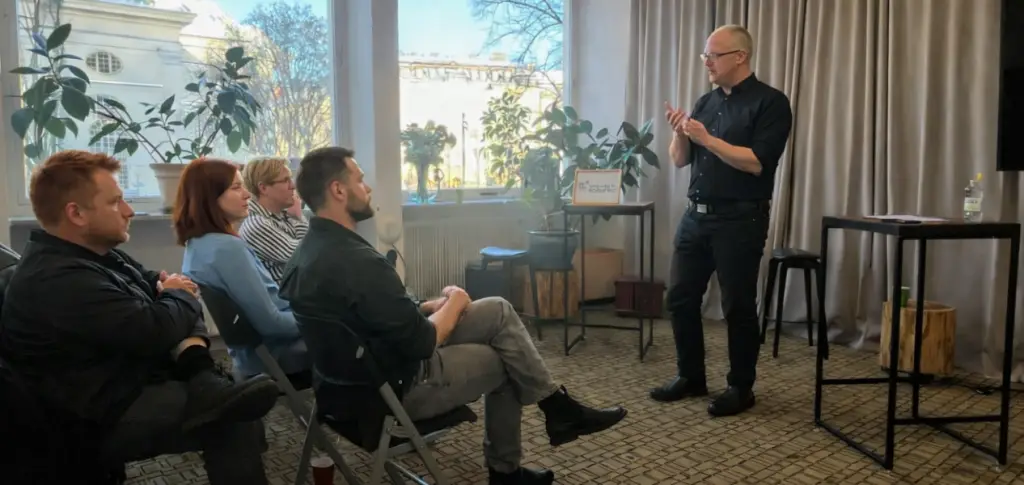 Hur ser ett tryggt fundament i talarträning ut i praktiken? En manlig kursledare står längst fram i ett ljust kursrum med stora fönster och beige gardiner. Han är klädd i mörk skjorta och mörka byxor, står upprätt med lätt framåtlutad hållning och håller händerna framför sig i en samlad gest som om han förklarar något steg för steg. Framför honom sitter fyra kursdeltagare på stolar i halvcirkel, vända mot honom och lyssnar koncentrerat. Utanför fönstret syns träd och en ljus byggnad, och i rummet finns gröna växter, ett litet bord och en hög pall bredvid honom.