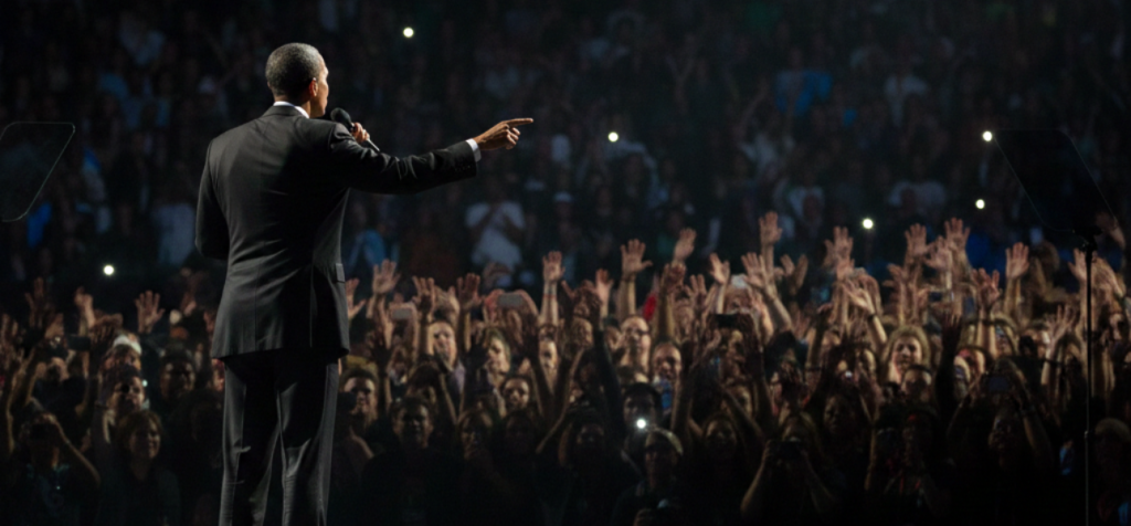 otografi taget bakifrån en talare på en mörk scen i en stor arena. Talaren är en mörkhyad man i mörk kostym som håller en mikrofon och pekar ut mot en enorm publik. Publikhavet är suddigt men man ser tusentals uppsträckta händer och små ljuspunkter från mobiltelefoner som skapar en elektrisk atmosfär."