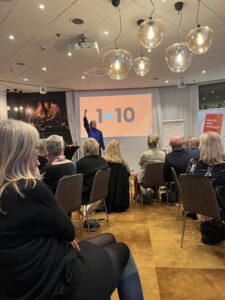 Hur kan du enklare interagera med din publik genom frågor och skalor? En person står längst fram i ett rum med sittande publik och håller upp ena armen högt i luften. Personen bär en blå tröja och vänder sig mot publiken med en öppen, engagerande hållning. Framför personen syns en projicerad bild med siffrorna ”1–10” i stort format. Publiken består av vuxna personer som sitter tätt på stolar i rader, vända mot scenen. Rummet är upplyst med varma lampor i taket och har karaktären av ett mindre föreläsnings- eller mötesrum.