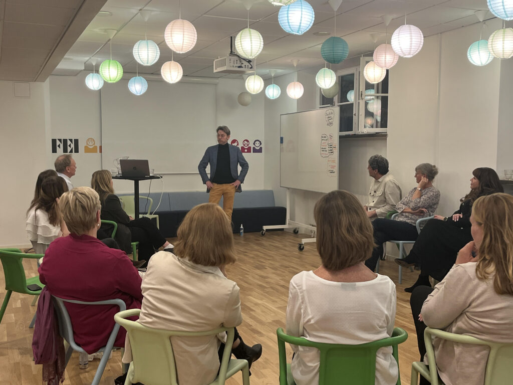 En man står i mitten av ett rum med trägolv och talar till en grupp människor som sitter i en cirkel på färgglada stolar. Han bär en mörk kavaj och ljusa byxor och har händerna placerade vid höfterna i en stabil, öppen hållning. Runt honom sitter deltagare i olika åldrar, sedda bakifrån och från sidan, med kropparna vända mot honom i ett lyssnande fokus. I taket hänger flera runda papperslampor i olika färger som ger rummet ett mjukt, informellt ljus. Bakom talaren syns whiteboardtavlor och ett mindre podium, vilket ger intryck av en workshop- eller utbildningsmiljö.