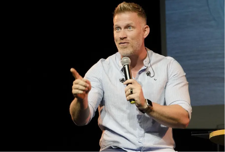 En man står på en mörk scen och talar i en handhållen mikrofon. Han är ljushyad, har kort ljust hår och lätt skäggstubb. Han bär en ljusblå långärmad skjorta med uppkavlade ärmar. Med ena handen håller han mikrofonen nära munnen, med den andra pekar han framåt i en tydlig gest. Hans hållning är upprätt och engagerad, och ansiktsuttrycket är fokuserat och intensivt, riktat mot publiken.