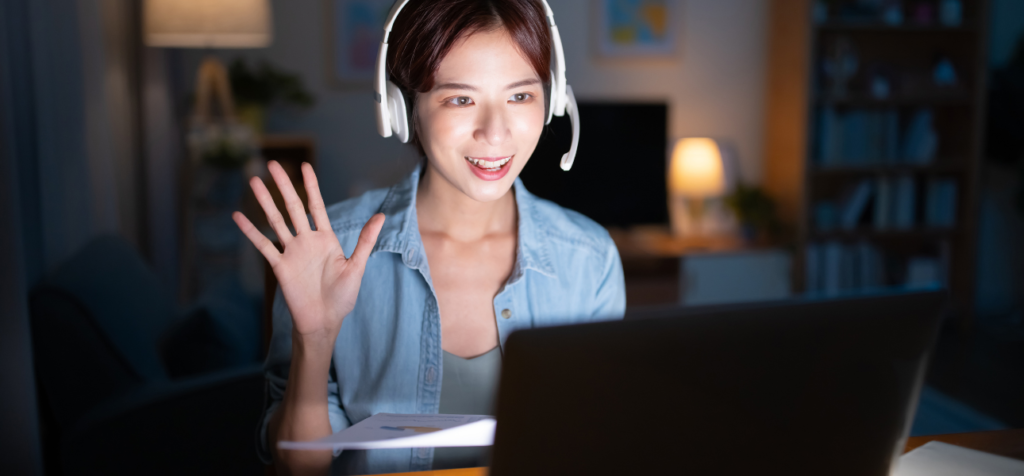 En kvinna med headset sitter framför en bärbar dator i ett kvällsbelyst rum. Hon ler, tittar in i kameran och vinkar med ena handen, som i ett hälsande eller inbjudande tilltal. Bilden förmedlar digital närvaro, dialog och ett aktivt möte mellan människor via skärm.