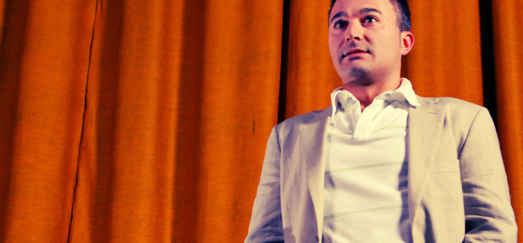 En man står på en scen framför en tung, orangefärgad ridå. Han är ljushyad, har kort mörkt hår och ett spänt men fokuserat ansiktsuttryck. Han bär en ljus kavaj över en vit pikétröja. Hans axlar är lätt uppdragna, armarna hänger nära kroppen och händerna är delvis spända, som i ett ögonblick av nervositet. Han står upprätt och ser rakt fram, som om han just ska börja tala inför publik.