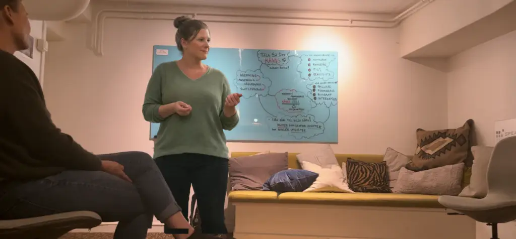 Hur skapar du en helhetsupplevelse i en presentation? En kvinna i 30–40-årsåldern står i ett mindre kursrum och talar till en sittande deltagare. Hon har uppsatt mörkblont hår i knut, grön stickad tröja och mörka jeans. Hennes händer är lätt upplyfta framför kroppen, fingrarna böjda som om hon förklarar något steg för steg. Bakom henne sitter en blå whiteboard med handritade cirklar, pilar och stödord. Rummet är varmt och informellt med soffa, kuddar och en stol vid sidan, vilket ger en workshop-känsla snarare än en föreläsningssal.