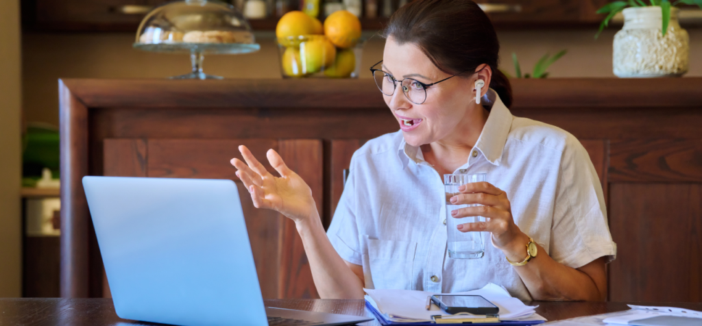 En kvinna med glasögon och håret uppsatt sitter vid ett bord framför en bärbar dator. Hon bär en ljus skjorta och trådlösa hörlurar, gestikulerar med ena handen och håller ett glas vatten i den andra. Hon ser engagerad ut och deltar aktivt i ett digitalt möte i en hemmiljö.