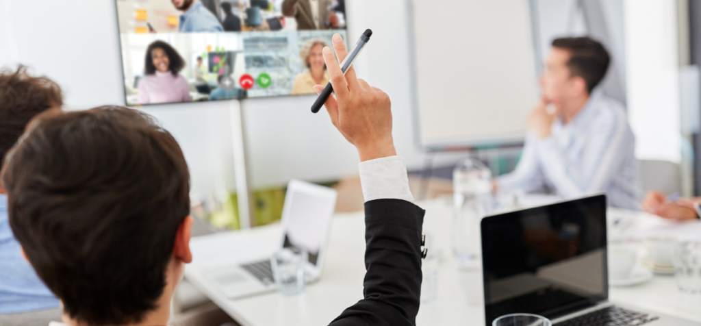 En person sitter i ett konferensrum och håller upp en penna för att bidra i samtalet. Runt bordet pågår ett fysiskt möte, samtidigt som flera deltagare deltar via en digital skärm. Bilden visar ett hybridmöte där närvaro och dialog delas mellan rummet och distans.