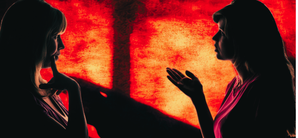 vad händer när vi väcker angelägenhet i vårt sätt att kommunicera? Två kvinnor står vända mot varandra i halvprofil mot en starkt röd och orange bakgrund som liknar upplysta fönster eller glödande ljus. Kvinnan till vänster har ljust hår och håller ena handen mot hakan i en eftertänksam gest. Kvinnan till höger har mörkare hår och håller ena handen upp med öppen handflata, som om hon förklarar eller ställer en fråga. Båda syns som mörka silhuetter mot ljuset, vilket gör deras kroppshållning och handrörelser tydliga. Bilden förmedlar ett intensivt och fokuserat samtal mellan två personer.