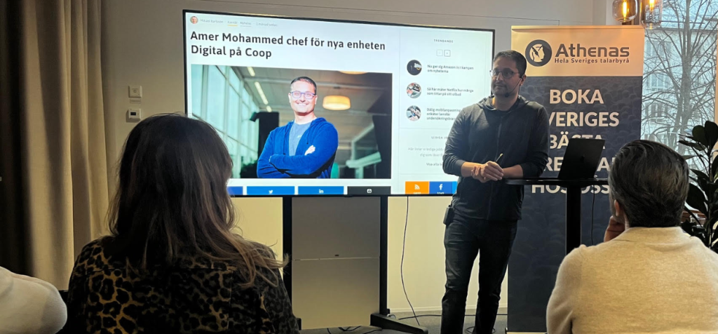 Hur skapar en föreläsare förtroende genom att ligga steget före sin publik? En man står längst fram i ett rum och håller i en mikrofon medan han talar till en sittande publik. Han bär mörka kläder och står stadigt med båda fötterna i golvet. Bakom honom syns en stor skärm med en presentation där hans porträtt och namn visas tillsammans med text om ledarskap och affärsutveckling. Publiken sitter tätt på stolar och är vänd mot honom, flera med uppmärksam hållning och blicken riktad framåt. Rummet har karaktären av ett modernt föreläsnings- eller mötesrum.