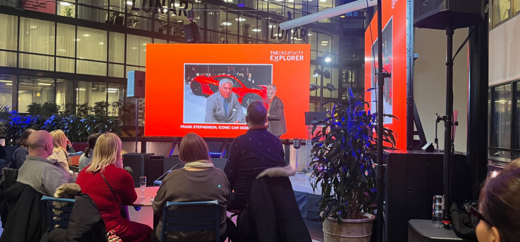 Hur visar en föreläsning att nyfikenhet är startpunkten för kreativitet? En man står på en scen i ett stort glasat atrium och talar till en sittande publik vid runda bord. Han håller kroppen lätt framåtlutad mot åhörarna i en aktiv berättande hållning. Bakom honom syns en stor röd presentationsskärm med en bild av en röd sportbil och en person på bilden, vilket fungerar som visuellt stöd till historien han berättar. Publiken sitter nära scenen, flera med huvudet vänt upp mot talaren och skärmen. Miljön liknar ett kvällsevent eller en inspirationsföreläsning med scenljus, högt i tak och växter runt scenområdet.