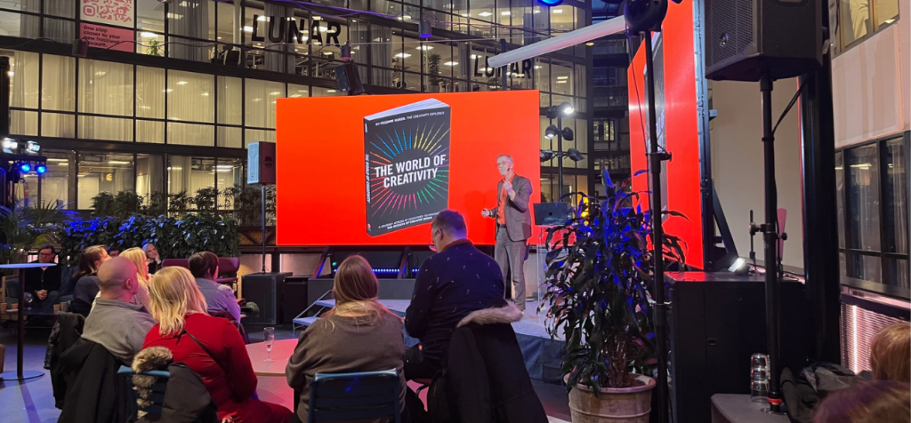 Hur kan kreativitet väckas genom att engagera publiken i en föreläsning? En man står på en scen i ett modernt, öppet atrium och föreläser inför en sittande publik. Han bär mörka kläder och står vänd mot åhörarna med en aktiv, framåtriktad hållning. Bakom honom syns en stor röd presentationsskärm med text och grafik som fungerar som visuellt stöd. Publiken sitter på stolar i rader, flera lutar sig lätt framåt och riktar blicken mot scenen. Rummet är högt i tak med glasväggar, ljus från omgivningen och en tydlig event- eller konferensmiljö.