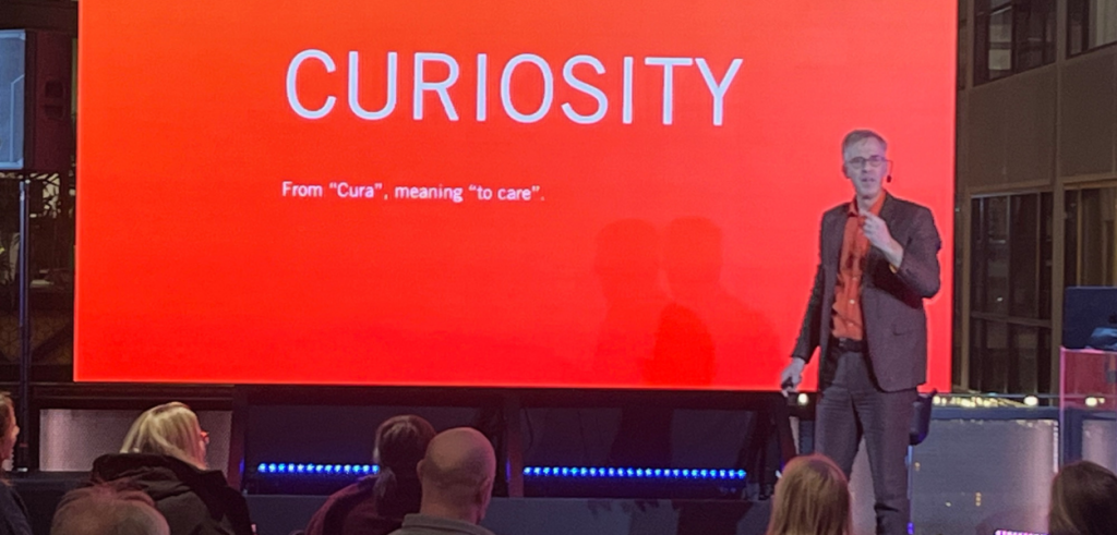 Hur kan en talare väcka nyfikenhet hos publiken genom ett tydligt budskap? En man står på en scen och håller en mikrofon i ena handen medan han talar till en sittande publik framför honom. Han bär kavaj, skjorta och glasögon och står vänd mot åhörarna med en rak men avslappnad hållning. Bakom honom täcker en stor röd presentationsskärm nästan hela väggen och visar ordet “CURIOSITY” i stora vita bokstäver samt en kort förklaring av ordets ursprung. Publiken sitter nedanför scenen med huvuden vända upp mot honom och skärmen, vilket skapar en tydlig fokuspunkt i rummet.
