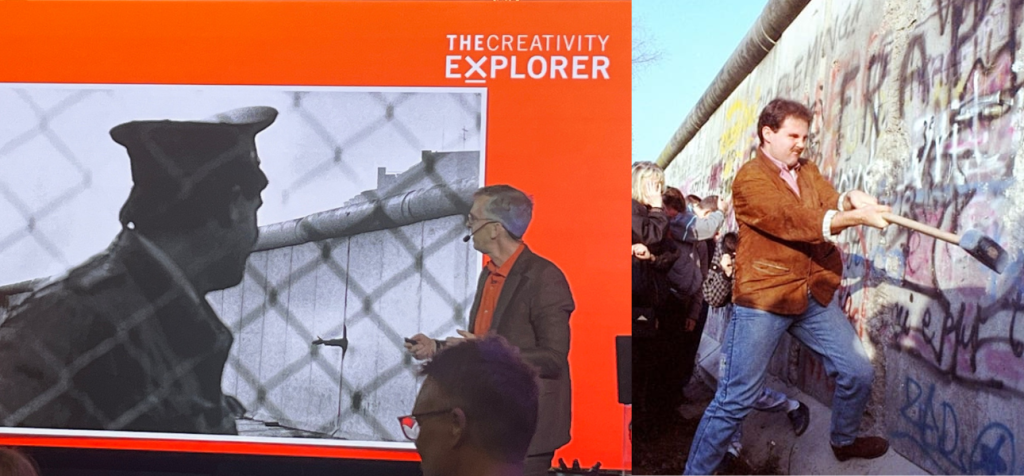 Hur kan ett perspektivskifte förändra hur publiken förstår kreativitet? En föreläsare står på en scen och pekar mot en stor presentationsskärm bakom sig. Han bär kavaj och glasögon och står lätt vriden mot bilden medan han förklarar för en sittande publik. På skärmen syns ett svartvitt fotografi av en soldat vid Berlinmuren bakom taggtråd. Bredvid visas ett historiskt färgfoto av en man som hackar i Berlinmuren med en slägga. Rummet är ett modernt atrium med högt i tak och publiken sitter framför scenen och tittar upp mot både talaren och bilderna.
