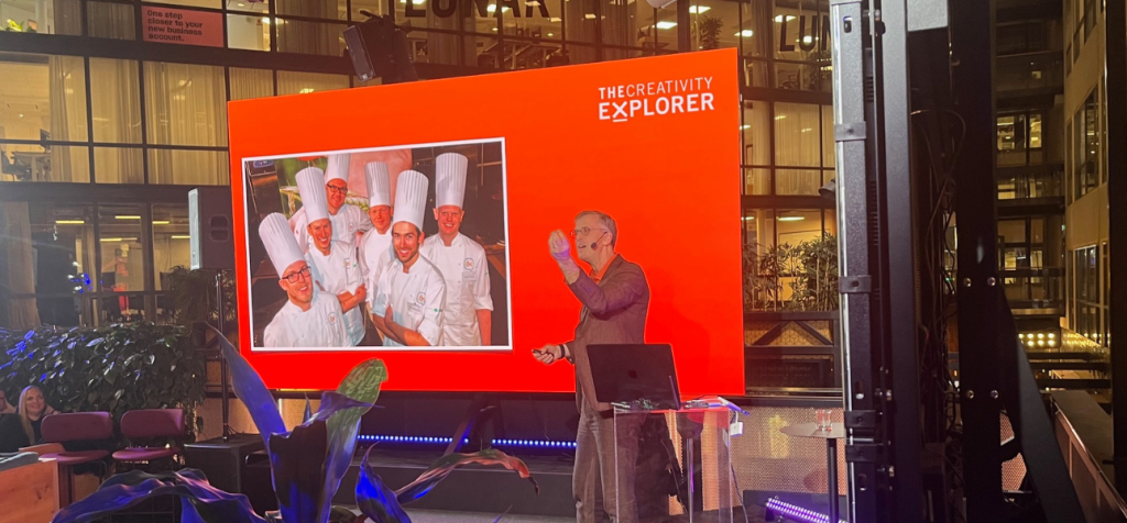 Hur kan en talare förklara kreativitet genom metaforer och igenkänning? En man står på en scen och föreläser med en mikrofon i handen medan han gestikulerar lätt med den andra handen i en förklarande rörelse. Han bär mörka kläder och är vänd mot en sittande publik. Bakom honom syns en stor röd presentationsskärm där en bild av flera kockar i vita kockmössor visas, vilket används som visuell metafor i föreläsningen. Publiken sitter nedanför scenen och tittar upp mot honom och skärmen i ett modernt glasat atrium med högt i tak, kvällsbelysning och växter i rummet.