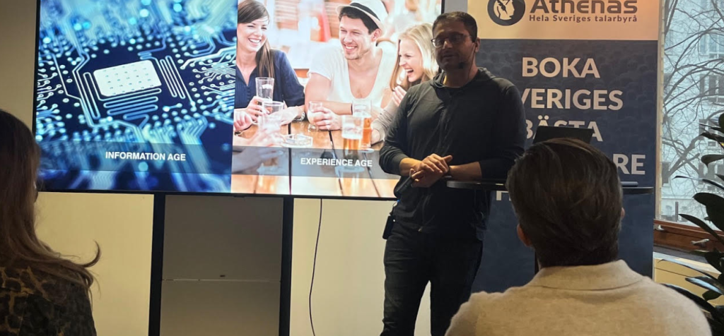 Varför är igenkänning avgörande för att retoriska grepp ska landa hos publiken? En man står längst fram i ett rum och föreläser för en sittande publik. Han bär mörka kläder och håller händerna samlade framför kroppen i en lugn, kontrollerad gest. Bakom honom syns en stor presentationsskärm uppdelad i två bildytor: till vänster ett närbildsliknande motiv av teknik och kretsar, till höger människor i samtal runt ett bord. Publiken sitter tätt och är vänd mot talaren. Miljön liknar ett modernt mötes- eller föreläsningsrum där visuella liknelser används för att skapa förståelse och igenkänning.