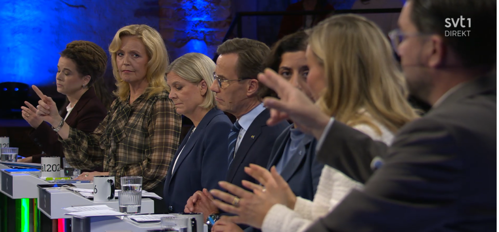 Hur påverkas en politisk debatt när frågor, svar och kroppsspråk konkurrerar om uppmärksamheten? Flera politiker sitter på rad under en direktsänd tv-debatt. I förgrunden gestikulerar två personer med händerna medan andra deltagare tittar mot dem, ned mot bordet eller åt sidan. Ansiktsuttrycken varierar från koncentrerade och allvarliga till avvaktande. Framför deltagarna ligger papper och står glas med vatten, och i bakgrunden syns blå scenbelysning.